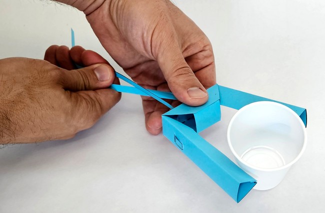 Hands hold an origami paper robot gripper, which grabs a small plastic cup