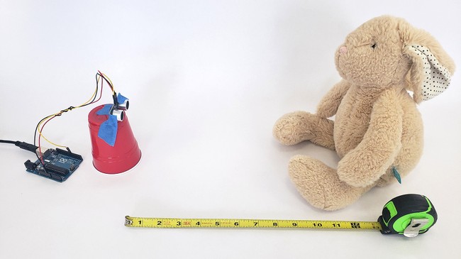 An Arduino connected to an ultrasonic sensor that is mounted on a cup, with a tape measure showing the distanced to a stuffed bunny rabbit