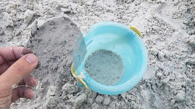 A circular piece of window screen held above a bucket. The bucket has a small layer of sand in the bottom.
