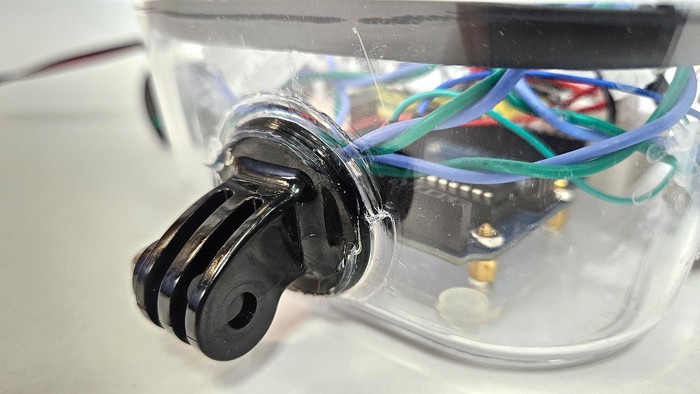 A camera mounting bracket bolted to the front of a plastic container with clear silicone sealant around the edge 