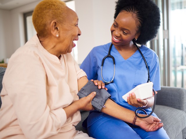 A nurse assistant in scrubs checks the blood pressure of an older woman, both seated indoors and smiling at each other.