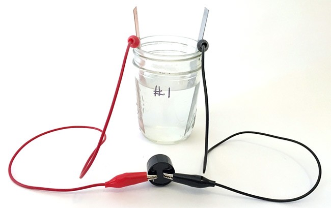 Alligator clips attach leads of a buzzer to a copper strip and zinc strip which both rest in a glass filled with saltwater