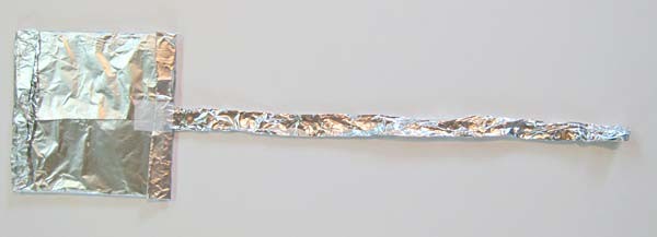 A long strip of aluminum foil is taped to the back of a cardboard square that is wrapped in foil