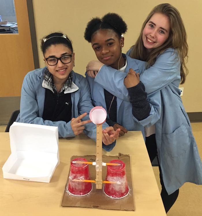 Three students next to a ball launcher made from plastic cups, pencils, a ruler and tape