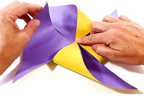 Hands folding cut paper square into a pinwheel.