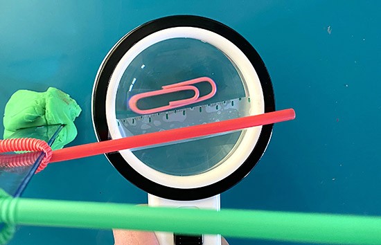 Top down view, looking through a lens held at the level of the straw down to a paperclip. A ruler that is stuck on the lens allows to measure the length of the paperclip is it is seen in the lens.
