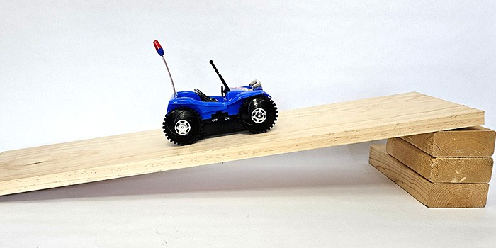 A battery-powered toy car drives up a ramp made from a board propped up with blocks at one end.