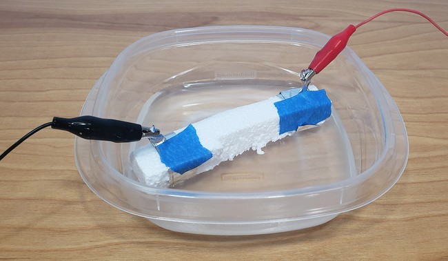 The foam conductivity sensor floating in a plastic food storage container of water, with alligator clips attached to the aluminum foil at each end