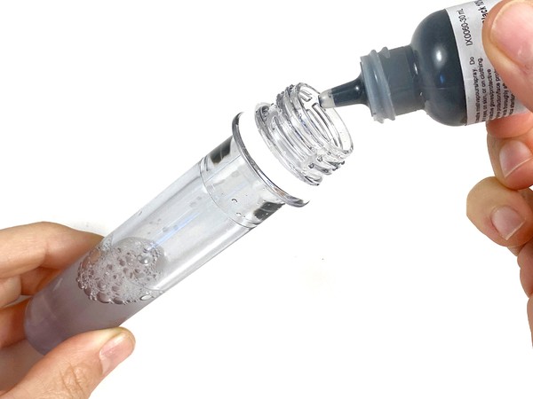 A hand holds a test tube filled with liquid. Another hand holds a small bottle of black ink above the test tube.