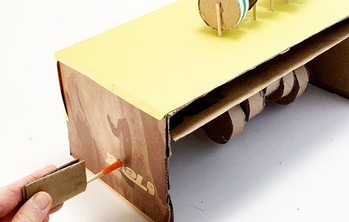 A side view of the automaton where a long section of the axle is sticking out. A piece of cardboard is being glued to this end of the axle.