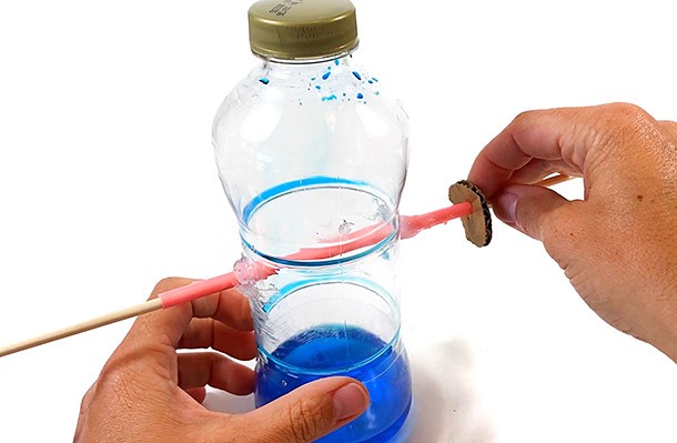 A plastic bottle containing some colored water with a straw sticking through two holes in the sides of the bottle. A wooden skewer is inserted through the straw.