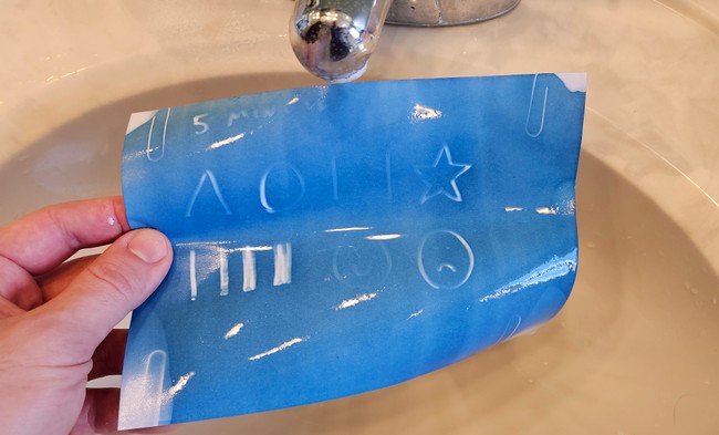 Sun print paper is rinsed under a faucet, revealing a blue background with white designs.