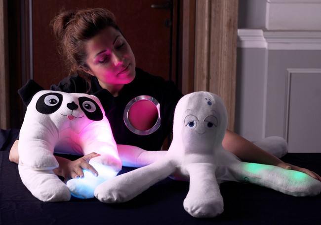 A woman sits next to two robots, a dog and an octopus, that are soft and cute. They are covered in fabric and stuffing, like plush toys.