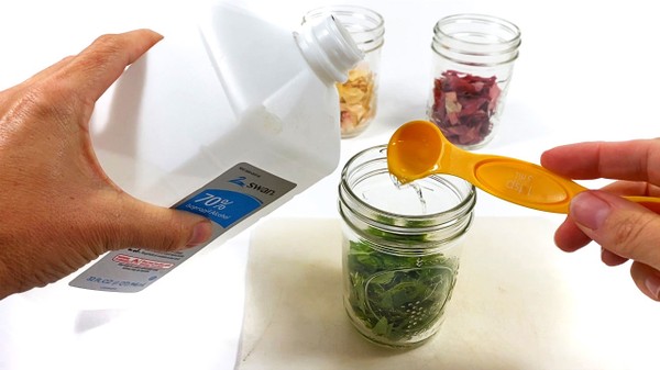 A mason jar filled with cut green leaves stands on a table. A teaspoon is held above the jar. The teaspoon is filled with 70% isopropyl alcohol.