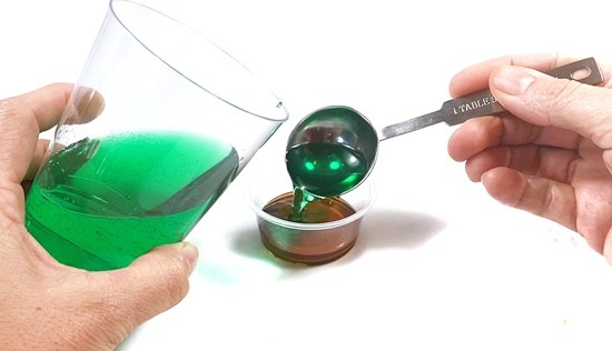 A mini cup with dark corn syrup in it is standing next to a cup with green water. A tablespoon full of green water is poured into the mini cup.