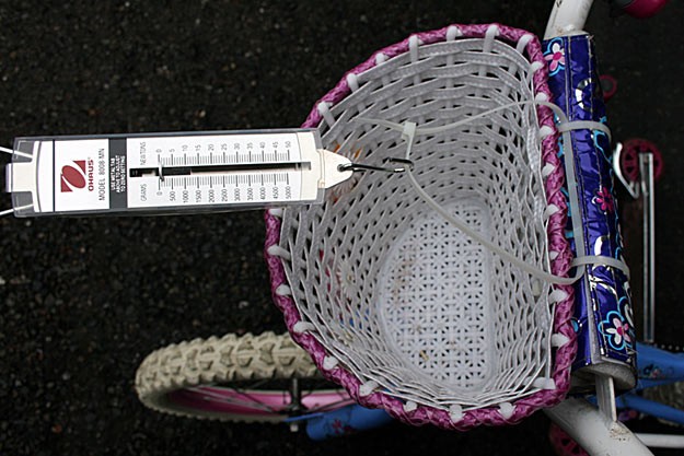 A spring scale is attached to the center of a bikes handlebars using plastic zip ties