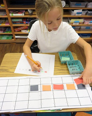 A student aligns small squares of paper into a grid that is drawn on a separate sheet of paper