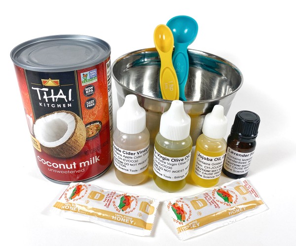 A can of coconut milk, several small bottles filled with liquids, 2 packets of honey, and a bowl with two measuring spoons on a table.