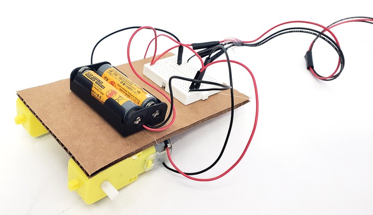 A piece of cardboard with the battery pack and breadboard glued to the top, and the motors glued to the bottom
