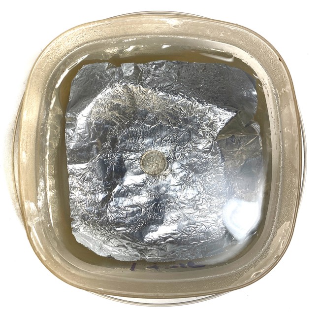 Top view of a plastic container lined with a sheet of aluminum foil. The container is filled with saltwater and a silver round lies on top of the aluminum foil. 