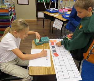 Two students align small squares of paper into a grid that is drawn on a separate sheet of paper