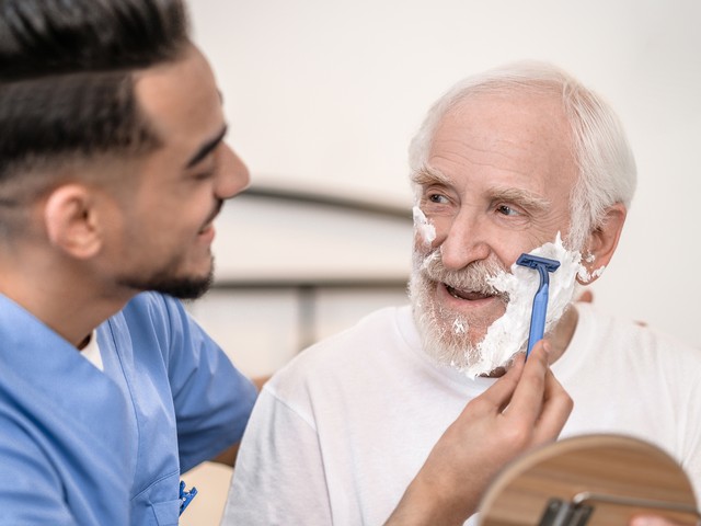  A nurse assistant carefully shaves the face of an older man.