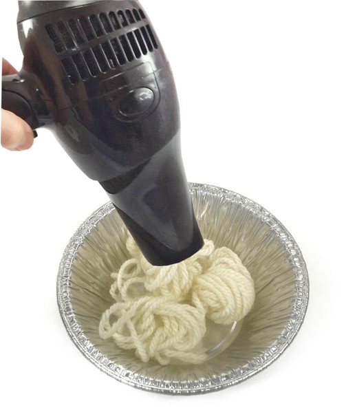 A hand holding a blow dryer on top of a bowl. Inside the bowl is a piece of wool yarn.