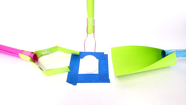 A grabber made from paper and string grasping a ping pong ball.