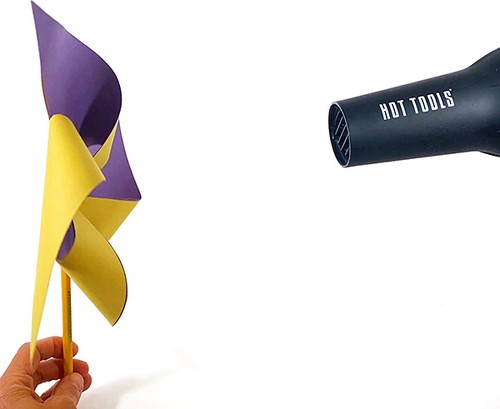 One hand holding a pinwheel. Another hand holding a blow dryer that is directed towards the pinwheel.