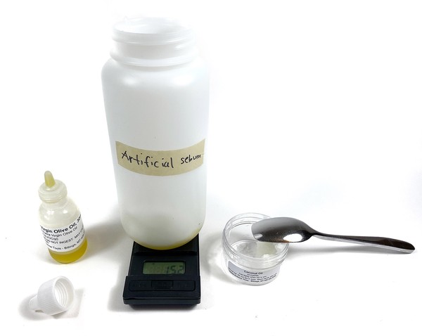 A small bottle filled with liquid and an cup with a spoon are standing next to a digital scale. On the scale stands a large bottle filled with some liquid.