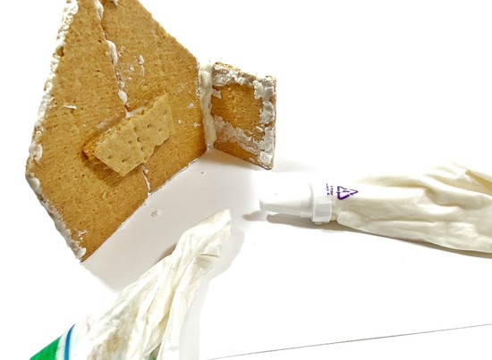 A partially built Graham cracker house with two bags of royal icing next to it. 