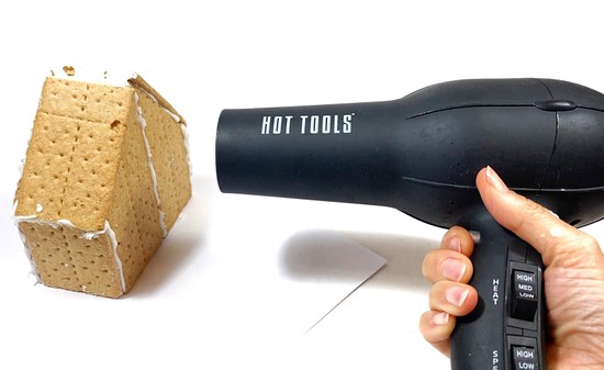 A Graham cracker house stands on a table. A hand holds a hairdryer about 10 cm away from the house, pointing it on one of the house's walls. 