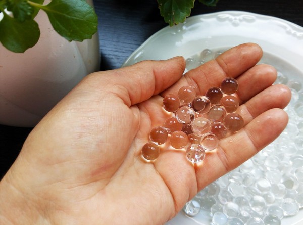 A hand holds colorless, transparent water gel beads. Each bead is approximately one centimeter in diameter.