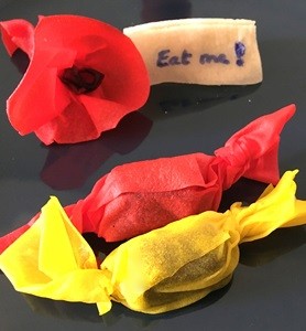 A flour from red edible paper, a name tag with the words 'Eat me' and two candies wrapped in red and yellow edible paper.