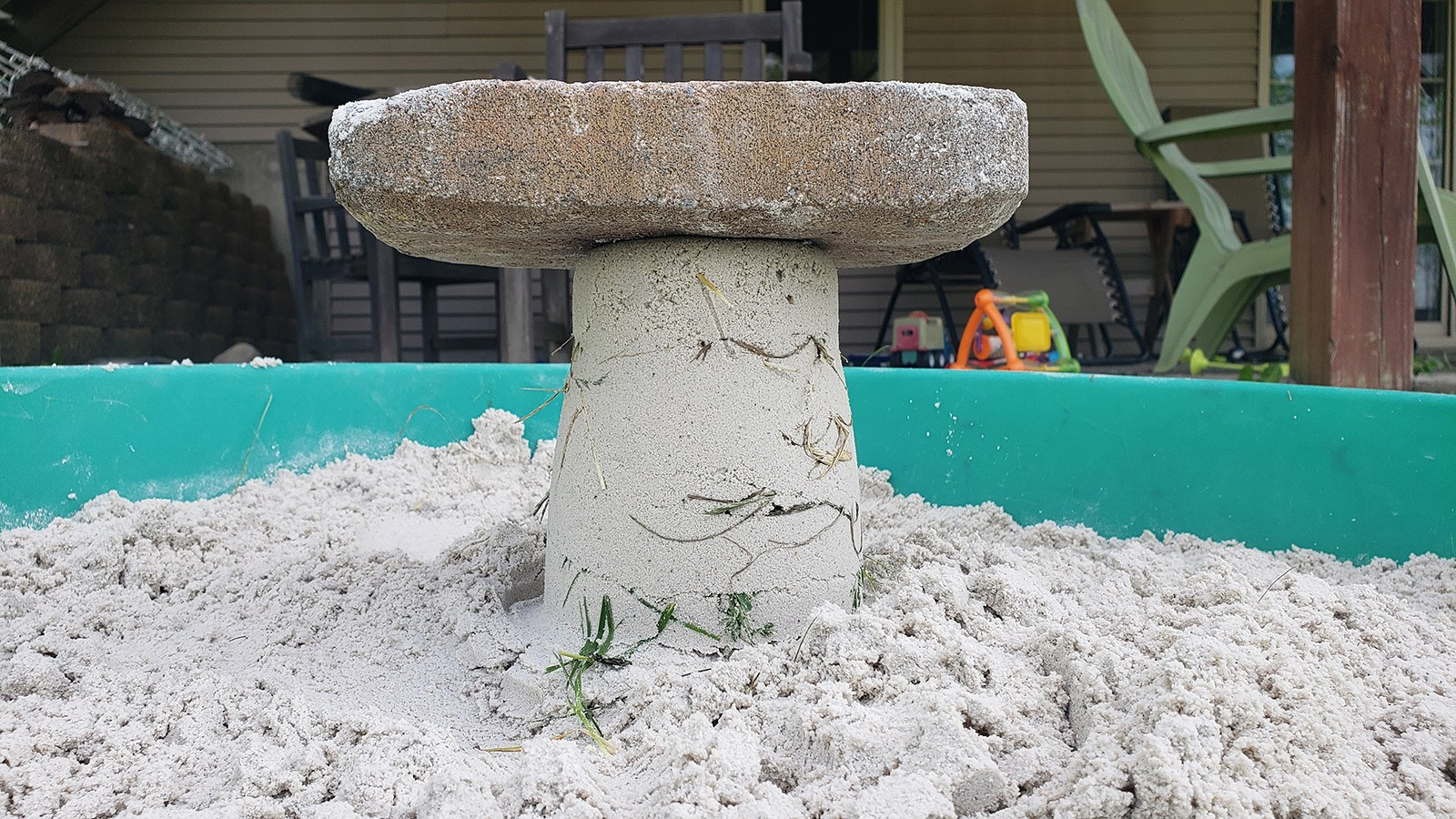 A bucket-shaped sandcastle in a sandbox with some pieces of grass sticking out the sides and a brick resting on top.