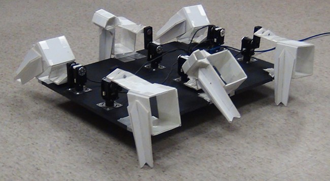 A robot with a flat body and six motorized legs made from folded foamboard. 