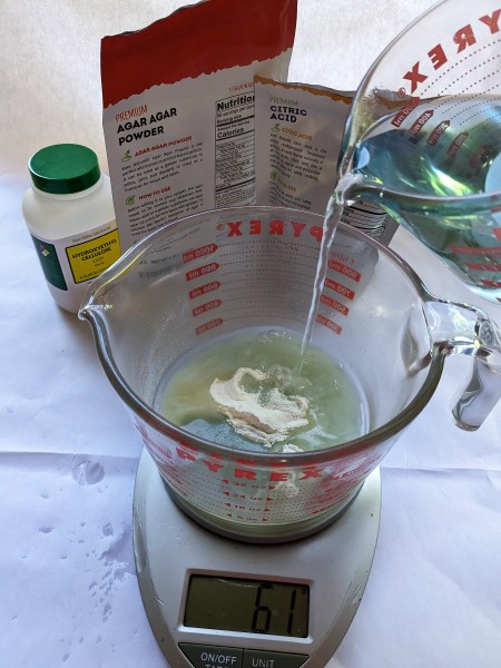 A pyrex measuring cup containing agar powder sitting on a scale while boiling water is poured into it to make a hydrogel.
