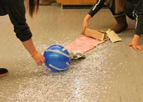 A photograph shows two students holding a balloon and piece of cardboard over scattered piles of shredded paper on the floor.