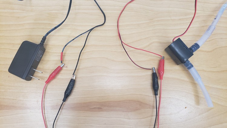 A DC wall adapter with two wires connected to a pump with two wires using two alligator clip leads. Red and black leads are used to indicate positive and negative, respectively.