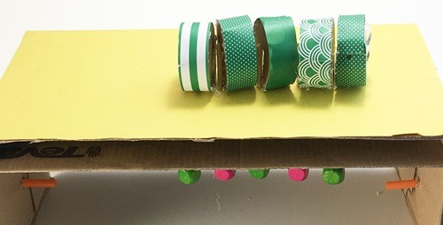 A cardboard box with five short cylinders glued to sticks sitting on top. The box has a second cardboard panel. 5 beads are visible below the second panel, one under each body part.