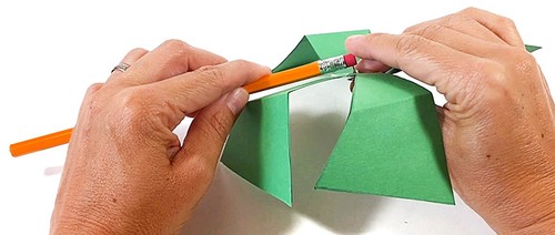 Hands holding a paper pinwheel made from 4 paper triangles. The paper pinwheel is attached to the pencil eraser with a thumbtack.