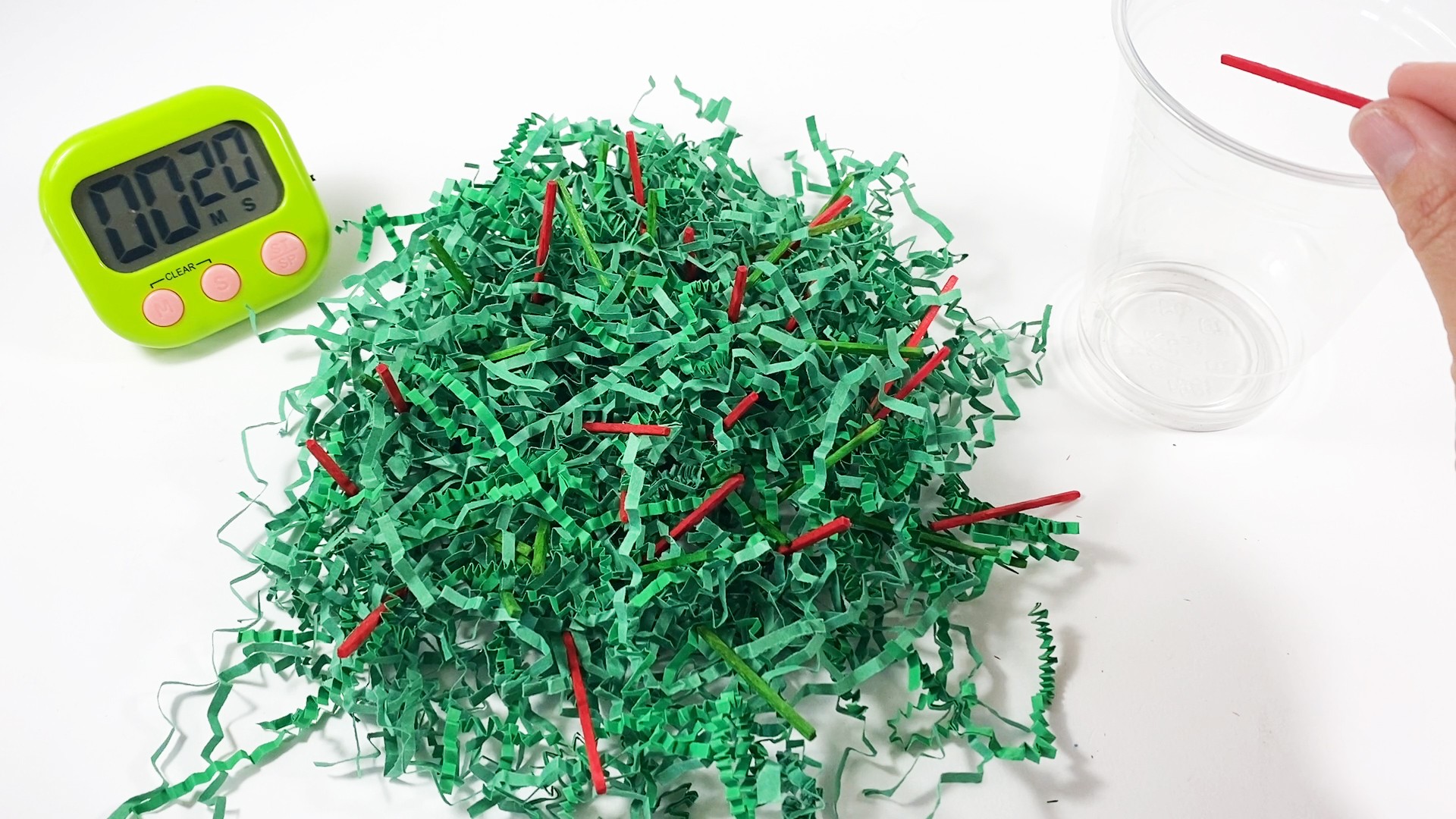 Green paper shreds with green and red matchsticks inside. A timer shows 20 seconds. A hand places a red stick into a cup.