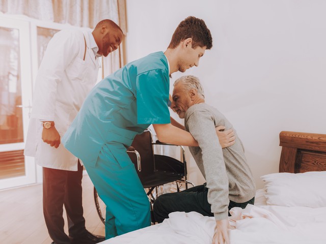 A nurse assistant helps to lift an older man off a bed and helps him into a wheelchair.