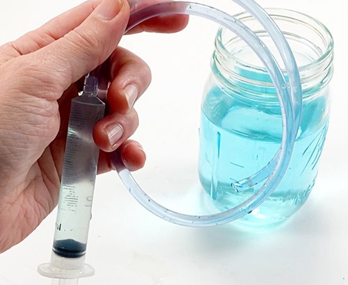 A small syringe connected to a piece of tube. Both the tube and the syringe are full of water. A jar of water is visible in the background.