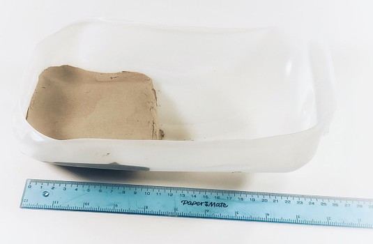Side view: A gallon jug cut in half, lengthwise, in which a clay platform is formed in the area farthest away from the spout. A ruler indicates the pavement is about 10 cm long.