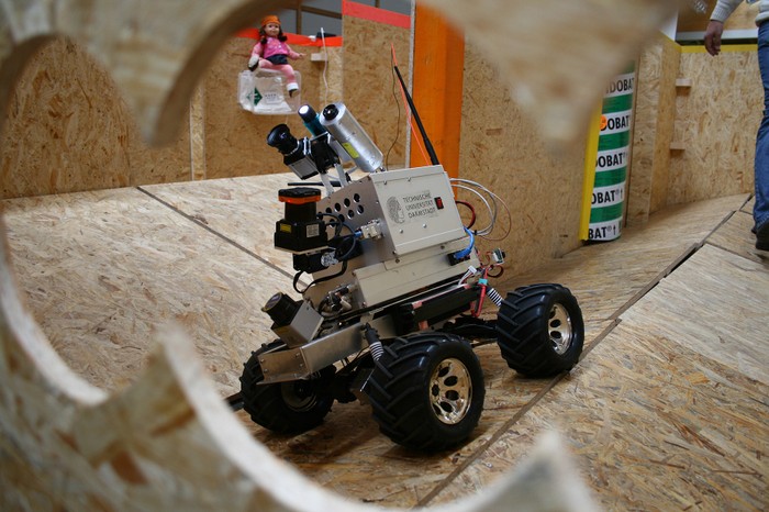 An all-terrain robot drives around an uneven plywood environment. A child-sized doll represents a survivor.