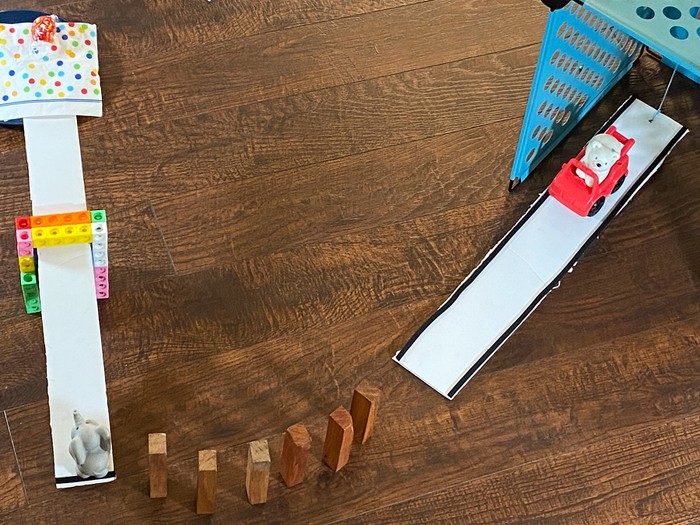 A rube goldberg machine with a car on a ramp aimed at several dominoes which are pointed toward a toy elephant on a see saw.