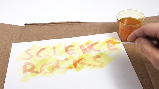 Hand holding a Q-tip soaked with turmeric solution and painting it over the paper with the secret message.
