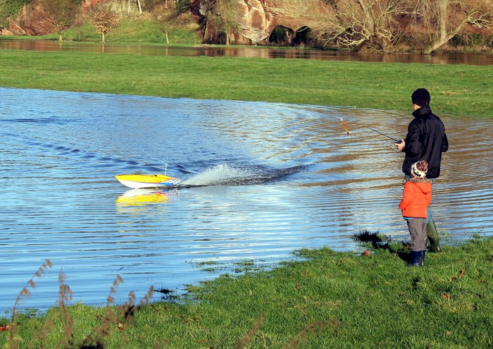 An adult and a child standing next to a body of water drive a boat using a remote control.
