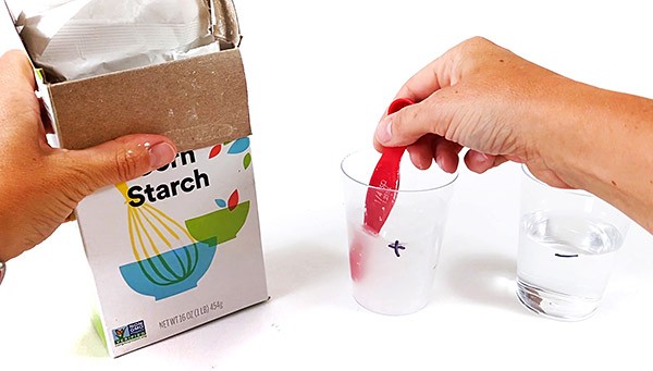 Hand holding a teaspoon and stirring a solution inside a cup. Another hand holds a box of cornstarch next to the cup.
